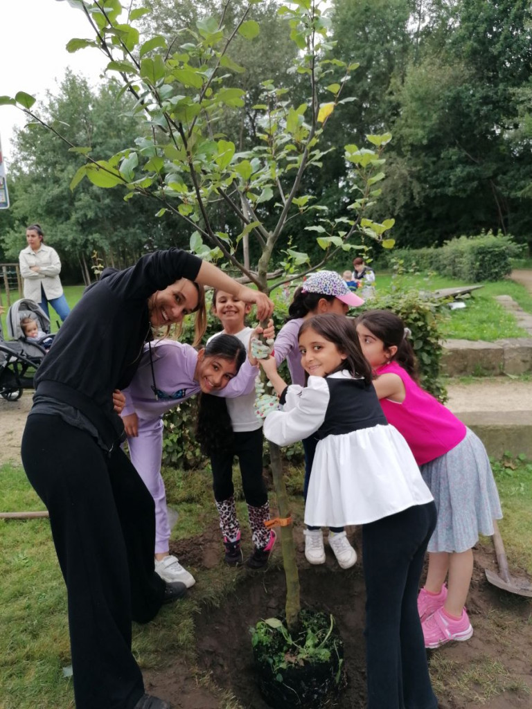 sechs Kinder und eine Betreuerin halten den frisch gepflanzten Apfelbaum am Stamm und strahlen Richtung Kamera