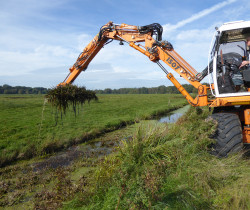 Bagger, der neben einem Graben im Grünland steht und tropfende, grüne Vegetation aus dem Graben hebt.