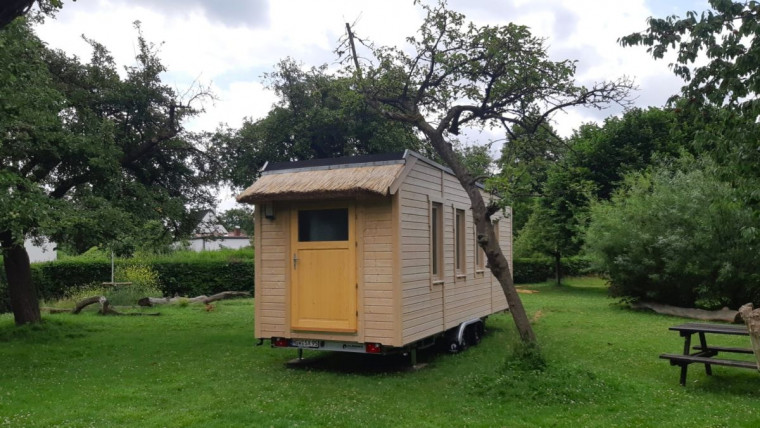 Holzwagen auf grüner Wiese zwischen Obstbäumen. Die Tür geht nach hinten raus und ist mit einem Schilfdach geschützt. 