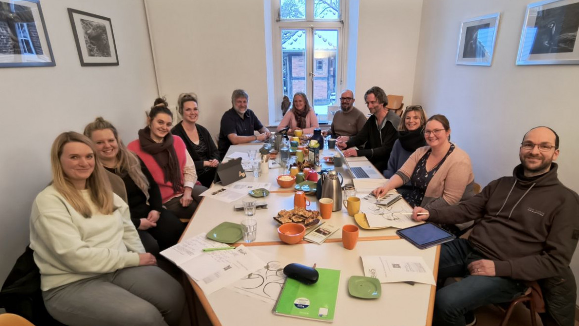 Elf Menschen sitzen in einem Besrpechungsraum und grinsen Richtung Kamera. Im Hinergund ist ein Fenster aus dem ein SChneebedecktes kleines Fachwerkhaus zusehen ist. Auf dem Tisch stehen Kaffee und Kuchen und es liegen viele Notizzettel rum. An den Wänden links und Rechts hängen je 2 Bilder.