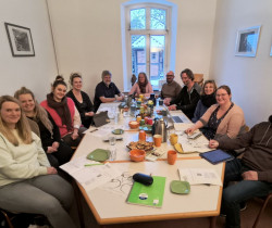 Elf Menschen sitzen in einem Besrpechungsraum und grinsen Richtung Kamera. Im Hinergund ist ein Fenster aus dem ein SChneebedecktes kleines Fachwerkhaus zusehen ist. Auf dem Tisch stehen Kaffee und Kuchen und es liegen viele Notizzettel rum. An den Wänden links und Rechts hängen je 2 Bilder.