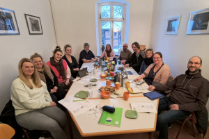 Elf Menschen sitzen in einem Besrpechungsraum und grinsen Richtung Kamera. Im Hinergund ist ein Fenster aus dem ein SChneebedecktes kleines Fachwerkhaus zusehen ist. Auf dem Tisch stehen Kaffee und Kuchen und es liegen viele Notizzettel rum. An den Wänden links und Rechts hängen je 2 Bilder.